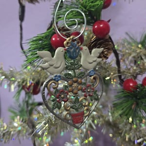 May include: A Christmas tree ornament featuring two white doves above a heart-shaped floral design. The ornament has a silver-colored frame and is decorated with red, blue, and brown accents. The ornament is hanging on a Christmas tree.