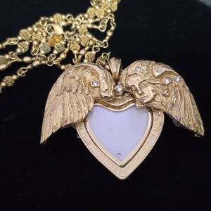 Puede incluir: Un collar con colgante en forma de coraz&oacute;n dorado. El colgante presenta dos figuras de &aacute;ngeles con alas, enmarcando un espacio blanco en forma de coraz&oacute;n. Peque&ntilde;as piedras transparentes est&aacute;n engastadas en las alas y las caras. La cadena del collar tambi&eacute;n es dorada.