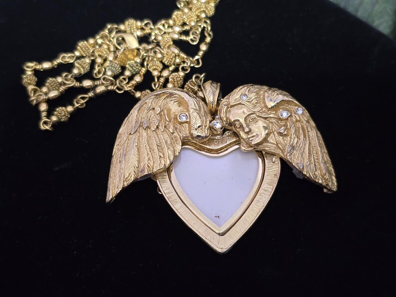 Puede incluir: Un collar con colgante en forma de coraz&oacute;n dorado. El colgante presenta dos figuras de &aacute;ngeles con alas, enmarcando un espacio blanco en forma de coraz&oacute;n. Peque&ntilde;as piedras transparentes est&aacute;n engastadas en las alas y las caras. La cadena del collar tambi&eacute;n es dorada.