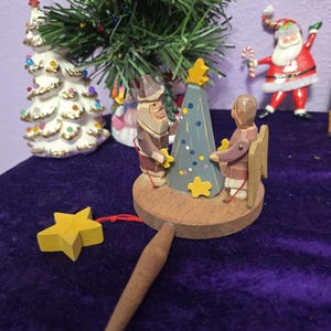 May include: A wooden mechanical toy featuring two carved figures standing on a circular base around a blue triangular tree. The figures are connected by red strings to yellow wooden stars, with a wooden handle extending from the base.