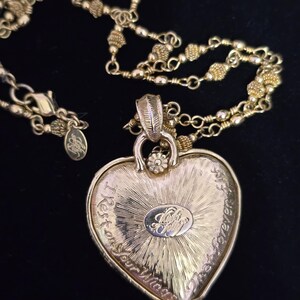 Puede incluir: Collar con colgante de coraz&oacute;n dorado con una cadena delicada. El colgante en forma de coraz&oacute;n tiene texto grabado y un emblema central. El collar est&aacute; sobre un fondo negro, resaltando los detalles de la joya.