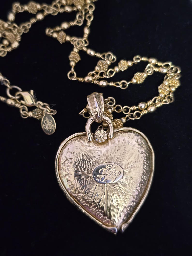Puede incluir: Collar con colgante de coraz&oacute;n dorado con una cadena delicada. El colgante en forma de coraz&oacute;n tiene texto grabado y un emblema central. El collar est&aacute; sobre un fondo negro, resaltando los detalles de la joya.