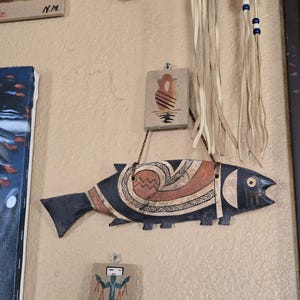 May include: A wooden fish-shaped wall hanging with a dark blue body and cream-colored accents. The fish features intricate geometric designs in brown and cream. Additional small, rectangular art pieces are also visible.