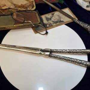 May include: A pair of silver-toned metal hair curling tongs featuring intricate embossed floral patterns on the handles. The tool consists of two long, slender, hinged rods that meet at a central pivot point, designed for styling hair with heat.