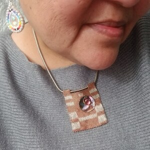 K&ouml;nnte beinhalten: Eine silberne Kettenkette mit einem quadratischen Anh&auml;nger aus gewebten braunen und wei&szlig;en Perlen. Der Anh&auml;nger hat eine kleine, runde, rosa und wei&szlig;e Muschel in der Mitte.