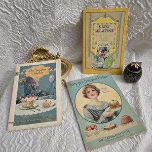 May include: A collection of vintage cookbooks and ephemera. The books feature colorful illustrations and text, including "Knox Gelatine" and "Sixty-five Delicious Dishes." A small, ornate black and gold container is also present.
