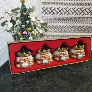 May include: A set of four ceramic owl figurines in a red box. Each owl has a white body, a gold crown, and a red and white striped candy cane. A small Christmas tree is in the background.