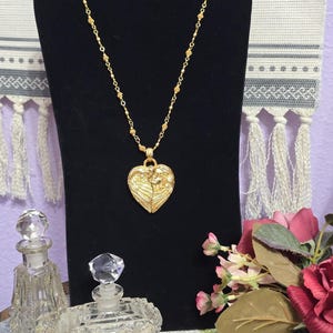 Puede incluir: Un collar dorado con un colgante en forma de coraz&oacute;n, exhibido sobre un busto de terciopelo negro. El colgante presenta un dise&ntilde;o en relieve. Dos frascos de perfume de vidrio y un ramo de flores rosas y rojas est&aacute;n en primer plano.