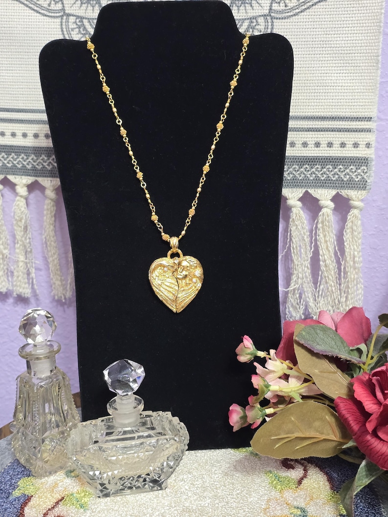 Puede incluir: Un collar dorado con un colgante en forma de coraz&oacute;n, exhibido sobre un busto de terciopelo negro. El colgante presenta un dise&ntilde;o en relieve. Dos frascos de perfume de vidrio y un ramo de flores rosas y rojas est&aacute;n en primer plano.