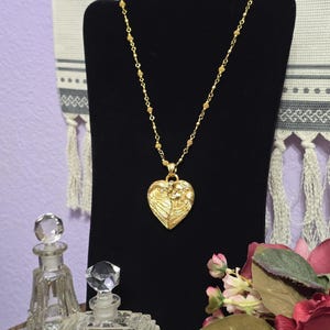 Puede incluir: Un collar dorado con un colgante en forma de coraz&oacute;n con un dise&ntilde;o de alas. El collar se exhibe en un soporte negro, con dos frascos de perfume de vidrio transparente en primer plano. El fondo incluye un arreglo floral y un textil decorativo.