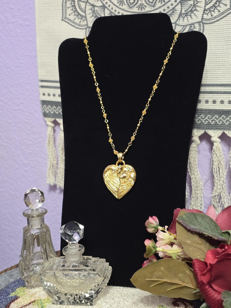 Puede incluir: Un collar dorado con un colgante en forma de coraz&oacute;n con un dise&ntilde;o de alas. El collar se exhibe en un soporte negro, con dos frascos de perfume de vidrio transparente en primer plano. El fondo incluye un arreglo floral y un textil decorativo.