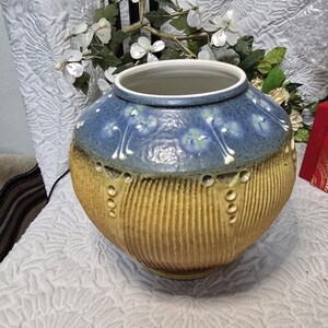 May include: A round ceramic vase featuring a textured yellow lower body with vertical ridges and a smooth blue upper section decorated with white and green floral motifs. The vase has a wide circular opening and a distinct transition between the two colors.