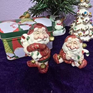 May include: Two ceramic Santa Claus figurines, one standing and one seated, with red suits and white beards. A green and red Christmas tin and a small Christmas tree are in the background. The figurines are on a purple surface.