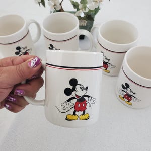 Op de afbeelding: Witte keramische mokken met Mickey Mouse. Elke mok heeft een zwarte en rode streep bij de rand en een Mickey Mouse-afbeelding. De mokken hebben een handvat en zijn geschikt voor warme of koude dranken.