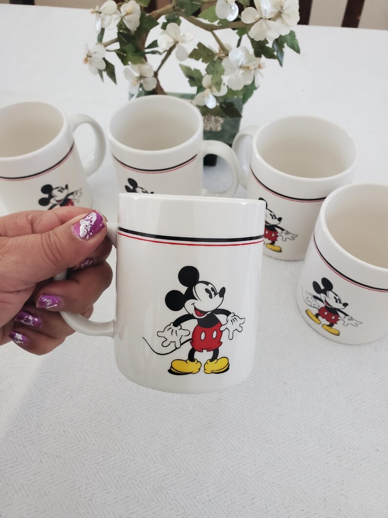 Op de afbeelding: Witte keramische mokken met Mickey Mouse. Elke mok heeft een zwarte en rode streep bij de rand en een Mickey Mouse-afbeelding. De mokken hebben een handvat en zijn geschikt voor warme of koude dranken.