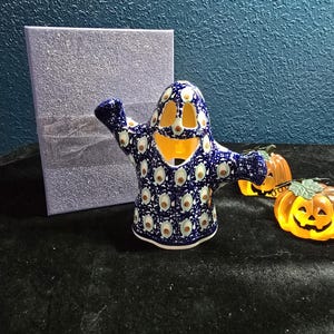 May include: A ceramic ghost figurine featuring a dark blue glaze with a repeating pattern of white and brown eye-like motifs. The ghost has cutouts for eyes and a mouth, with arms extended outward, and is positioned next to a silver gift box.