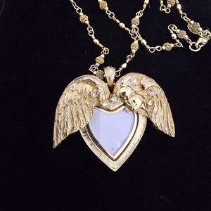 Puede incluir: Collar dorado con un colgante en forma de coraz&oacute;n con alas de &aacute;ngel y una cara. El colgante tiene un centro blanco y est&aacute; adornado con peque&ntilde;os cristales. La cadena es delicada y con cuentas, con un cierre de mosquet&oacute;n.