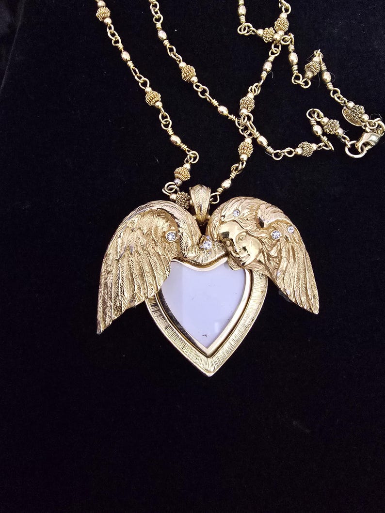 Puede incluir: Collar dorado con un colgante en forma de coraz&oacute;n con alas de &aacute;ngel y una cara. El colgante tiene un centro blanco y est&aacute; adornado con peque&ntilde;os cristales. La cadena es delicada y con cuentas, con un cierre de mosquet&oacute;n.