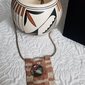 K&ouml;nnte beinhalten: Eine silberne Kettenkette mit einem Anh&auml;nger aus Kupfer- und wei&szlig;en Perlen, der eine runde, schillernde Muschel mit einem Kupferakzent zeigt.