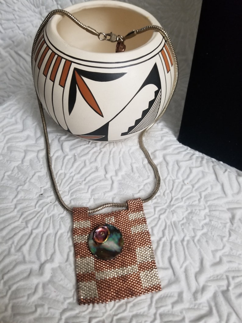K&ouml;nnte beinhalten: Eine silberne Kettenkette mit einem Anh&auml;nger aus Kupfer- und wei&szlig;en Perlen, der eine runde, schillernde Muschel mit einem Kupferakzent zeigt.