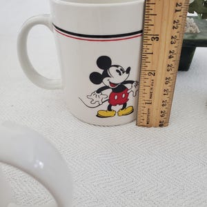 Op de afbeelding: Witte keramische mok met Mickey Mouse. De mok heeft een zwarte en rode streep bij de rand. De mok is ongeveer 10 cm hoog. Mickey Mouse is afgebeeld in zijn klassieke outfit met een rode broek en gele schoenen.