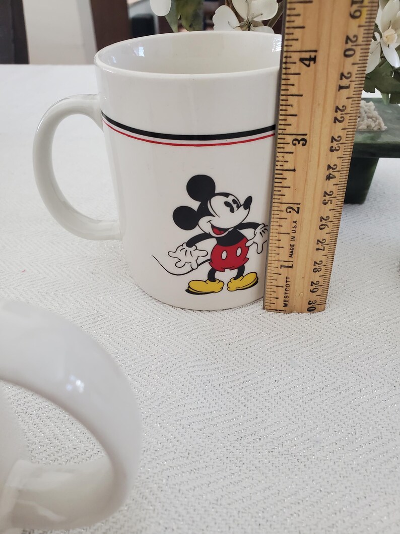 Op de afbeelding: Witte keramische mok met Mickey Mouse. De mok heeft een zwarte en rode streep bij de rand. De mok is ongeveer 10 cm hoog. Mickey Mouse is afgebeeld in zijn klassieke outfit met een rode broek en gele schoenen.