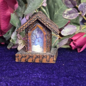 May include: A miniature wooden church with intricate carvings and a small window displaying a religious scene. The church is brown with a decorative roof and sits on a square base. The background includes flowers.