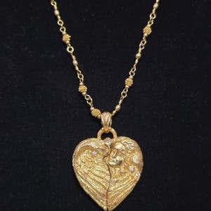 Puede incluir: Un collar dorado con un colgante en forma de coraz&oacute;n. El colgante presenta un dise&ntilde;o esculpido de un rostro con alas, adornado con peque&ntilde;as piedras brillantes. La cadena del collar es de color dorado delicado.