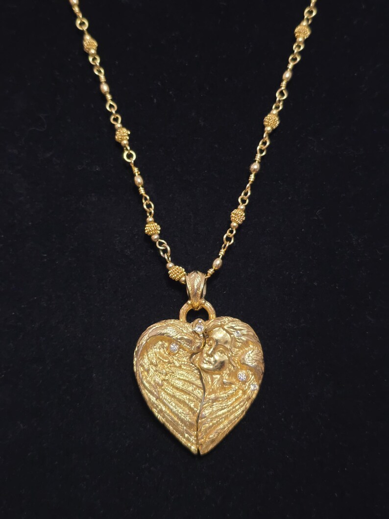 Puede incluir: Un collar dorado con un colgante en forma de coraz&oacute;n. El colgante presenta un dise&ntilde;o esculpido de un rostro con alas, adornado con peque&ntilde;as piedras brillantes. La cadena del collar es de color dorado delicado.