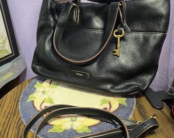 May include: A black leather handbag with two top handles and a detachable shoulder strap. The bag features a gold-tone key charm hanging from the handle and a rectangular metal plate with the word FOSSIL embossed on the front exterior panel.