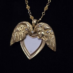 Puede incluir: Un collar con un medall&oacute;n en forma de coraz&oacute;n dorado, adornado con alas de &aacute;ngel y peque&ntilde;as piedras preciosas transparentes. El medall&oacute;n se abre para revelar un interior blanco. La cadena del collar tambi&eacute;n es dorada y tiene un dise&ntilde;o de cuentas.