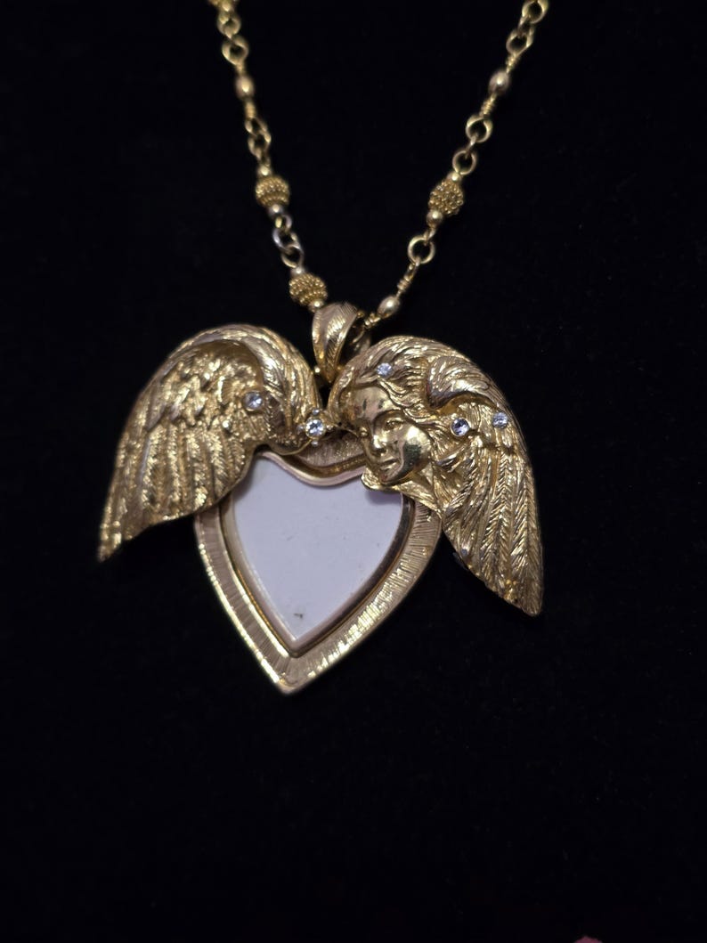 Puede incluir: Un collar con un medall&oacute;n en forma de coraz&oacute;n dorado, adornado con alas de &aacute;ngel y peque&ntilde;as piedras preciosas transparentes. El medall&oacute;n se abre para revelar un interior blanco. La cadena del collar tambi&eacute;n es dorada y tiene un dise&ntilde;o de cuentas.