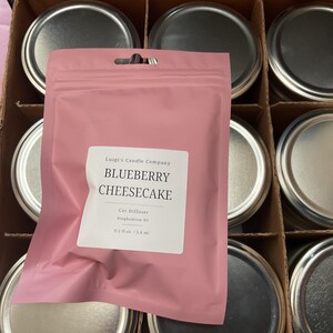 May include: A pink car air freshener bag with the text "BLUEBERRY CHEESECAKE" is displayed in a cardboard box with several metal-lidded jars. The bag is from Luigi's Candle Company.
