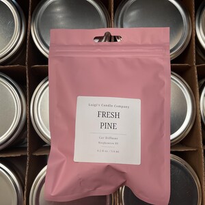以下が含まれることがあります： 「Fresh Pine」と「Luigi's Candle Company」の文字が白いラベルに書かれた、ピンク色の車用芳香剤バッグ。バッグは、銀色の蓋が付いた容器の前にあります。