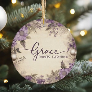 May include: Round Christmas ornament with a floral design and the words "Grace Changes Everything" in a cursive font. The ornament features purple flowers and greenery around the edge, with a beige background. It hangs from a natural twine.