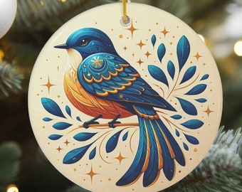 Könnte beinhalten: Ein runder Keramik-Anhänger mit einem blauen und orangefarbenen Vogel mit filigranen Details, der auf einem Zweig mit blauen Blättern sitzt. Der Hintergrund ist cremefarben mit goldenen Sternen.