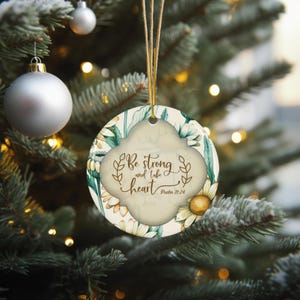 May include: Round Christmas ornament with a floral design and the text "Be strong and take heart Psalm 31:24". The ornament is cream-colored with a gold string for hanging. The background is a Christmas tree.