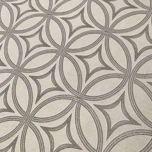 Op de afbeelding: Een witte stof met een herhalend patroon van overlappende cirkels en ruitvormige figuren in donkergrijs.