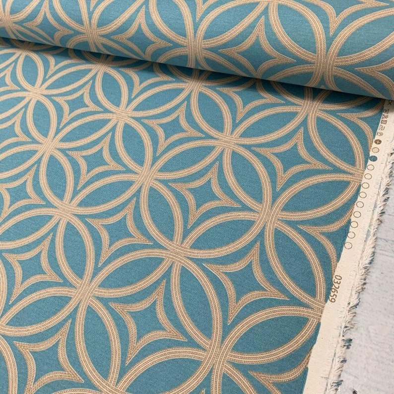 Op de afbeelding: Teal stof met een herhalend patroon van overlappende beige cirkels. De stof heeft een geweven textuur en is op zichzelf gevouwen. De rand van de stof is zichtbaar en heeft een witte rand met het nummer "032369" erop gedrukt.