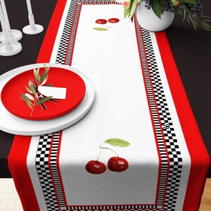 May include: A white table runner with a black and white checkered border and red cherry designs. The runner has a red border.