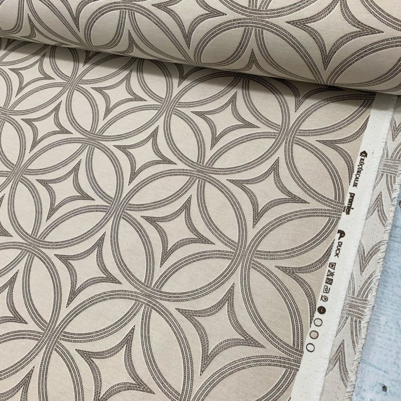 Op de afbeelding: Beige stof met een herhalend patroon van bruine, in elkaar grijpende cirkels en ruiten. De stof heeft een textuur en is gemarkeerd met "Duck Fabric" en "Remarkable Prints".
