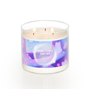 May include: A three-wick candle in a clear glass jar with a label that reads "late night gamer" and features a cartoon illustration of a computer screen, a keyboard, and a cute animal character. The candle is surrounded by a pink and blue glow.