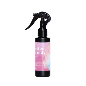 Può includere: Un flacone spray in plastica nera con un design di zucchero filato rosa e blu. La bottiglia è etichettata "cotton candy" ed è uno spray per ambienti.