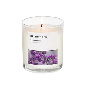 May include: A clear glass jar candle with a white label that reads "Heliotrope" and a floral image. The candle is lit and the flame is visible.