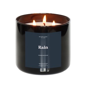 Op de afbeelding: Een zwarte glazen kaars met drie brandende lonten. De kaars heeft een donkerblauw etiket met het woord "Rain" en "Premium Wax Blend". De tekst "MENS COLLECTION" is ook zichtbaar. De kaars is gemaakt met essentiële oliën.