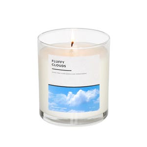 Puede incluir: Una vela encendida en un frasco de vidrio transparente. La vela tiene un interior de cera blanca y una etiqueta que dice "Fluffy Clouds" con un cielo azul y un gráfico de nubes. La vela es de Naladak Candles.