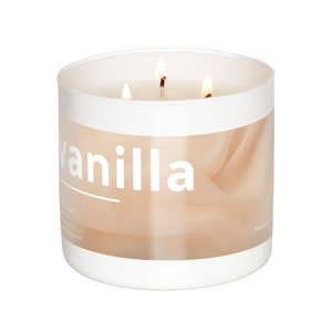 Op de afbeelding: Een witte kaars pot met een beige label waarop "vanilla" staat. De kaars heeft drie lonten en is aangestoken.