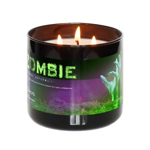 Puede incluir: Vela de tres mechas en un tarro de vidrio negro. La etiqueta verde dice "ZOMBIE" con un gráfico de mano de zombi verde. El aroma es una mezcla de pachulí y canela negra.