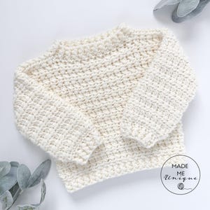 May include: A white crocheted sweater with a round neckline and long sleeves. The sweater is made with a textured stitch and has a relaxed fit.