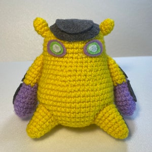 May include: A yellow crocheted plush toy with purple arms and a gray felt hat. The toy has green eyes with a white outline.